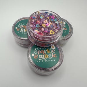 Puede incluir: Cuatro botes transparentes de purpurina para el cabello. La purpurina es una mezcla de colores y formas, incluyendo flores y corazones. Los botes tienen etiquetas verdes con el texto "it's all sparkle & magic HAIR GLITTER".