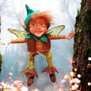 Puede incluir: Una muñeca de hada caprichosa con cabello rojo, pecas y ojos verdes. La muñeca lleva un sombrero verde, chaleco y leggings, con zapatos y alas marrones. Una pequeña mariquita roja decora un zapato. El hada está suspendida frente a un fondo borroso de bosque.