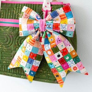 Può includere: Un colorato fiocco in tessuto patchwork, legato con un centro rosa, adorna una borsa tote verde con finiture rosa. Il fiocco è realizzato con piccoli pezzi di tessuto quadrati in vari colori e motivi.