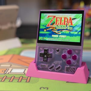 May include: A handheld gaming device with a screen displaying "The Legend of Zelda: The Minish Cap" game. The device has a transparent purple case with pink buttons and a pink stand. The game's title and copyright information are visible.