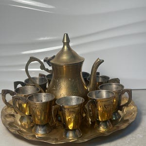 May include: A vintage brass tea set, including a teapot and several small cups, arranged on a matching tray. The set has an aged, antique appearance with intricate etched designs. The cups have handles and sit on a decorative tray.