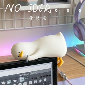 May include: A white duck-shaped desk accessory is perched on a computer monitor. The duck has a yellow beak and feet. The monitor displays a software interface. A headset with cat ears sits on a stand. The text "NO IDEA" and Chinese characters are visible.