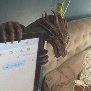 Front view of a dark bronze-effect 3D-printed dragon monitor buddy wrapped around the upper-right corner of a black computer screen. The fantasy dragon has a horned scaled head, open mouth, large claws gripping the top bezel and additional claws holding the side, with a blue wall and hanging green plants behind it.