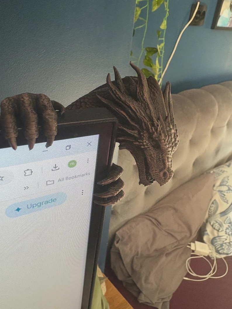 Front view of a dark bronze-effect 3D-printed dragon monitor buddy wrapped around the upper-right corner of a black computer screen. The fantasy dragon has a horned scaled head, open mouth, large claws gripping the top bezel and additional claws holding the side, with a blue wall and hanging green plants behind it.