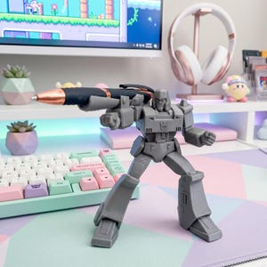 May include: A gray Megatron action figure holding a black and rose gold pen, posed as if aiming. The figure is on a desk with a pastel keyboard and a computer monitor in the background. The scene includes decorative succulents and headphones.