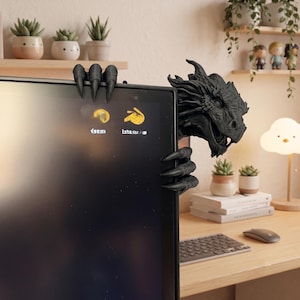 Front view of a dark bronze-effect 3D-printed dragon monitor buddy wrapped around the upper-right corner of a black computer screen. The fantasy dragon has a horned scaled head, open mouth, large claws gripping the top bezel and additional claws holding the side, with a blue wall and hanging green plants behind it.