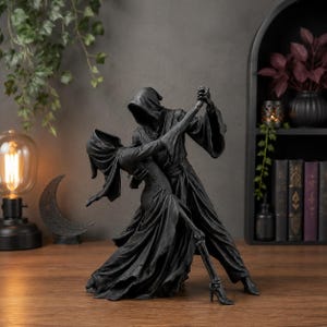 Könnte beinhalten: Eine schwarze Skulptur, die zwei kapuzentragende Figuren in einer Tanzpose darstellt. Eine Figur trägt ein langes, fließendes Gewand, während bei der anderen ein skelettiertes Bein mit einem hochhackigen Schuh sichtbar ist. Die Figuren tanzen.