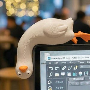 May include: A white goose-shaped laptop accessory with orange feet and beak. The goose is perched on top of a laptop screen. The screen displays the Autodesk 3ds Max 2021 software interface.