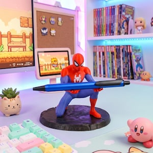 Puede incluir: Una figura de Spider-Man roja, azul y negra sosteniendo un bolígrafo azul. La figura está sobre una base marrón oscuro. También se ve una pequeña suculenta en una maceta con forma de gato y una figura de Kirby.