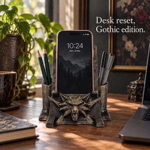 May include: A dark, weathered gold-toned desk organizer featuring a central smartphone stand and two side pen holders. The front is adorned with a sculpted wolf head emblem. The text "Desk reset, Gothic edition." is visible above the stand.