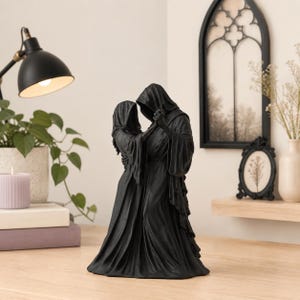 Könnte beinhalten: Eine mattschwarze Harzskulptur, die zwei kapuzenbedeckte Gestalten in fließenden Gewändern beim Tanzen darstellt. Die Figuren sind an Händen und Taille verbunden, ihre Gesichter sind unter tiefen Kapuzen verborgen, was eine dunkle, fließende Silhouette ergibt.