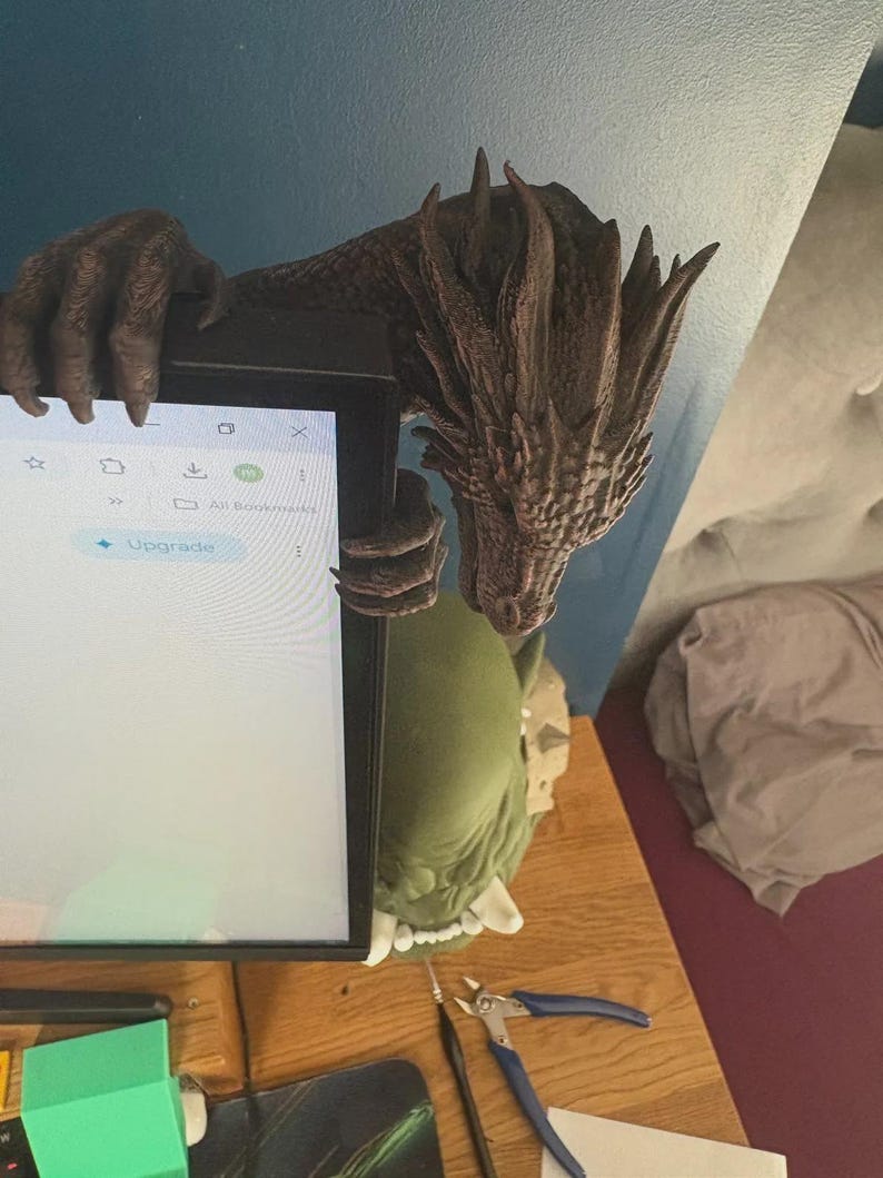 Front view of a dark bronze-effect 3D-printed dragon monitor buddy wrapped around the upper-right corner of a black computer screen. The fantasy dragon has a horned scaled head, open mouth, large claws gripping the top bezel and additional claws holding the side, with a blue wall and hanging green plants behind it.