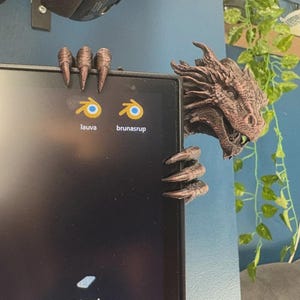 Puede incluir: Una figura de dragón texturizada de color marrón oscuro con garras afiladas que se agarran al borde superior y lateral de un monitor de computadora. La cabeza del dragón es visible a la derecha, con escamas detalladas, y sus garras sobre la pantalla.