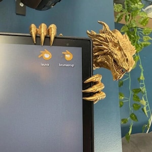 Front view of a dark bronze-effect 3D-printed dragon monitor buddy wrapped around the upper-right corner of a black computer screen. The fantasy dragon has a horned scaled head, open mouth, large claws gripping the top bezel and additional claws holding the side, with a blue wall and hanging green plants behind it.