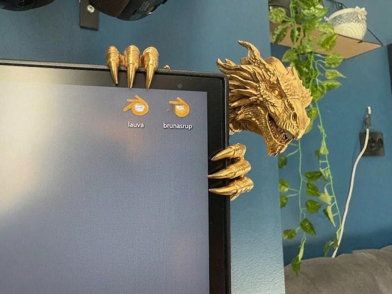 Front view of a dark bronze-effect 3D-printed dragon monitor buddy wrapped around the upper-right corner of a black computer screen. The fantasy dragon has a horned scaled head, open mouth, large claws gripping the top bezel and additional claws holding the side, with a blue wall and hanging green plants behind it.