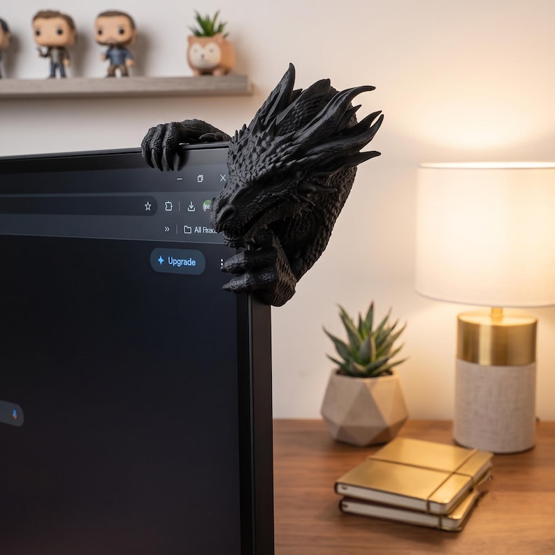 Dark bronze dragon monitor buddy displayed on the corner of a desktop computer in a gaming workspace. The highly detailed 3D-printed creature wraps around the monitor with its horned head on one side and claws gripping the top and front edges, making a striking gift for gamers and dragon collectors.