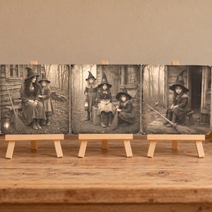 May include: Three square sepia-toned prints displayed on small wooden easels. The prints feature vintage-style photography of figures in pointed hats and dark clothing, posed with brooms and lanterns in a wooded setting near a rustic cabin.