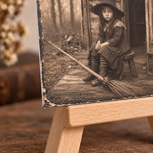 May include: A sepia-toned rectangular print displayed on a light-colored wooden easel. The image depicts a child wearing a wide-brimmed hat and dark clothing, sitting on a small stool on a wooden porch next to a broomstick with a forest background.