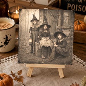 May include: A square sepia-toned print on a small wooden easel featuring three children dressed in pointed hats and dark clothing. One child holds a lit lantern, one sits on a wooden crate, and one holds a broomstick in front of a rustic building.