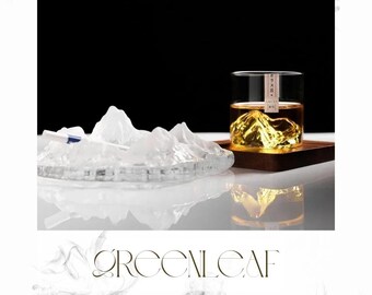 May include: A clear glass with a mountain-shaped ice cube and a wooden tray. A clear, mountain-shaped ice sculpture is in the foreground. The word "green" is at the top, and "GREENLEAF" is at the bottom. The image promotes nature-inspired home goods.