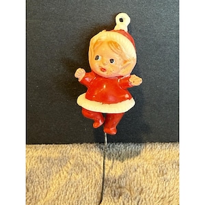 May include: A vintage Christmas ornament depicting a small child figurine in a red outfit with white trim. The figurine has a painted face and is attached to a metal wire. The ornament is likely from the mid-20th century.