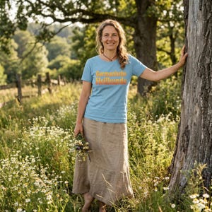 Puede incluir: Una mujer con una camiseta azul claro con el texto "Germanische Heilkunde" en naranja, y una falda color beige. Está en un campo de flores silvestres, sosteniendo un pequeño ramo. La escena es al aire libre, con un árbol y una valla de madera.