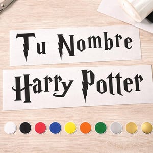 Op de afbeelding: Twee witte rechthoekige stickers met zwarte tekst, één met de tekst "Tu Nombre" en de andere met "Harry Potter" in een gestileerd lettertype. Er zijn ook een verscheidenheid aan gekleurde ronde stickers en een oranje schaar aanwezig.