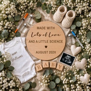 Op de afbeelding: Een witte babyromper met de tekst Baby Williams, een rond houten bord met de tekst Made with Lots of Love and a Little Science August 202X, twee injectiespuiten, twee glazen flesjes, beige gebreide babyslofjes, vier houten blokken met de letters BABY en een echo.