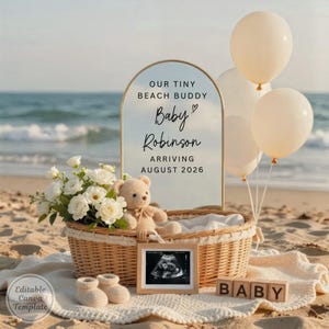 May include: A wicker basket containing a beige teddy bear, white flowers, and a framed ultrasound image. An arched sign reads: OUR TINY BEACH BUDDY Baby Robinson ARRIVING AUGUST 2026. Three cream balloons and wooden blocks spelling BABY sit nearby.