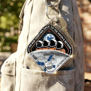 May include: Clear acrylic keychain with a space-themed design. The keychain features a diamond shape with the text "WE RISE TOGETHER, BACK TO THE MOON & BEYOND." The design includes astronauts, a planet, and a spacecraft. The keychain is attached to a backpack.