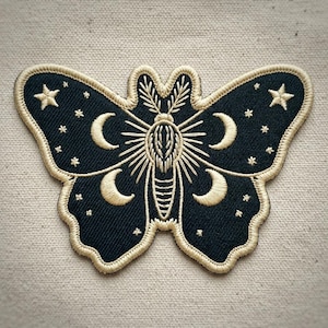 May include: Embroidered patch in the shape of a moth with a celestial design. The moth is navy blue with gold-colored embroidery. Features include crescent moons, stars, and a central sunburst design. The patch is approximately 4 inches wide.
