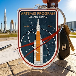 May include: Clear acrylic keychain featuring the Artemis Program logo with a rocket illustration. The keychain has a dark blue background with the text "ARTEMIS PROGRAM WE ARE GOING" and the NASA and ESA logos. Includes a leather strap and keys.