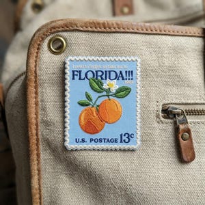 May include: A rectangular patch with a light blue background featuring a vintage Florida postage stamp design. The stamp depicts two oranges with green leaves and a white flower. Text includes "FLORIDA!!!" and "U.S. POSTAGE 13¢".