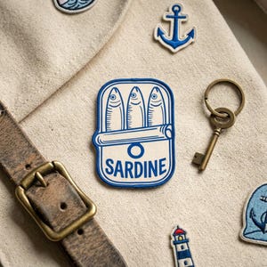 May include: A collection of nautical-themed patches and accessories. The central patch features a blue and white sardine can with the word "SARDINE". Other items include an anchor patch, a key, and a leather belt with a brass buckle.