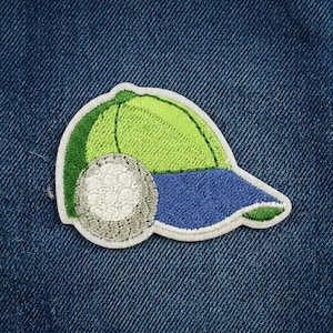 May include: Embroidered patch featuring a golf ball and a baseball cap. The cap is green and blue with a white border. The golf ball is white with gray detailing. The patch is designed to be ironed on or sewn onto fabric.