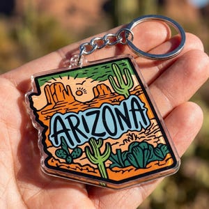 May include: A clear acrylic keychain shaped like the state of Arizona, featuring a colorful desert landscape illustration with orange rock formations, green cacti, a yellow sun, and the word ARIZONA printed in large, bold, light blue letters.