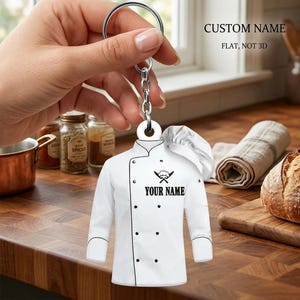 May include: A white chef's jacket keychain with black buttons and trim, a chef's hat, and the text "YOUR NAME" below crossed knives and a fork. The keychain is held by a silver chain and ring, with the text "CUSTOM NAME FLAT, NOT 3D" in the background.