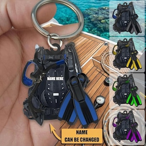 May include: A scuba diving-themed keychain featuring a black and blue diving apparatus, fins, and a metal keyring. The keychain has the text "NAME HERE" and "NAME CAN BE CHANGED". Additional color options of fins are shown in the background.
