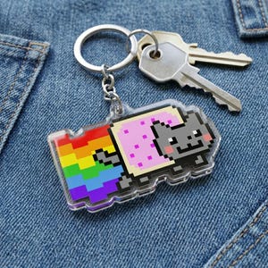 May include: A clear acrylic keychain featuring a pixelated Nyan Cat design. The cat has a gray body, pink pop-tart body, and a rainbow trail. The keychain is attached to a silver keyring with two keys.
