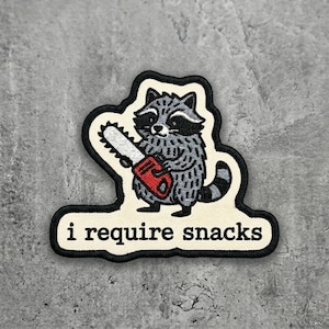 May include: Embroidered patch featuring a cartoon raccoon holding a red chainsaw. The raccoon is gray and white with a black mask and tail. The patch has the text "i require snacks" in a black font.