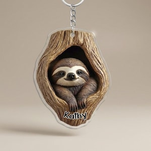May include: A keychain featuring a smiling sloth nestled in a tree trunk. The sloth is brown and white, with a friendly expression. The tree trunk is a light brown color. The name "Kathy" is printed in black script at the bottom.