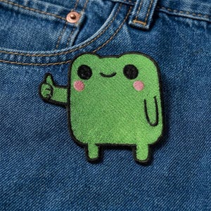 May include: Green embroidered frog patch with black eyes, a smiling mouth, and pink cheeks. The frog is giving a thumbs-up gesture. The patch is on a blue denim background.
