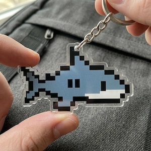 May include: A pixelated shark keychain with a silver chain and ring. The shark is light blue with black accents and two black eyes. The keychain is held against a gray background.