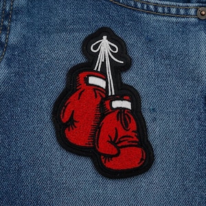 May include: Embroidered patch featuring two red boxing gloves with white accents, connected by white strings. The patch has a black border and is set against a blue denim background.