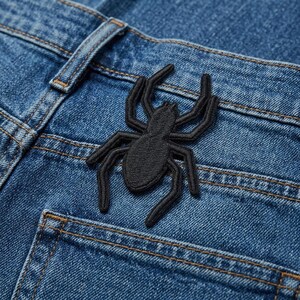 May include: A black embroidered spider patch is affixed to the back pocket of a pair of blue denim jeans. The spider has eight legs and a textured body. The jeans have visible stitching and a classic five-pocket design.