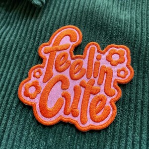 May include: An embroidered patch with the words "Feelin' Cute" in orange script on a pink background. The patch has an orange border and small flower accents. The patch is set against a green corduroy fabric.