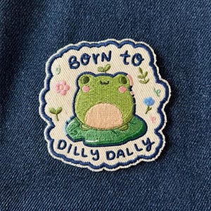 May include: Embroidered patch featuring a green frog sitting on a lily pad. The patch has a white border with the words "Born to Dilly Dally" in blue. Pink and blue floral accents surround the frog.