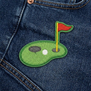 May include: A green embroidered patch featuring a golf course design. The patch includes a golf ball, a hole, and a red flag on a yellow pole. The patch is sewn onto a blue denim fabric.