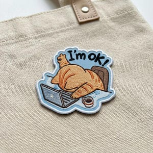 May include: Embroidered patch with a cartoon cat resting on a desk with a laptop and coffee cup. The patch has a light blue border and the words "I'm ok!" in black text.