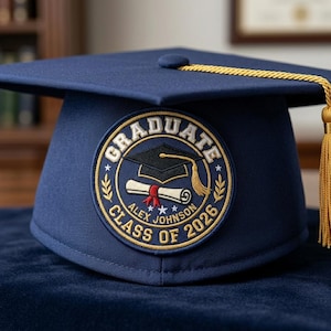 May include: A navy blue graduation cap with a gold tassel. The cap features a circular patch with the word "GRADUATE" and "CLASS OF 2026" in gold lettering. The patch also includes a mortarboard, diploma, and the name "ALEX JOHNSON".